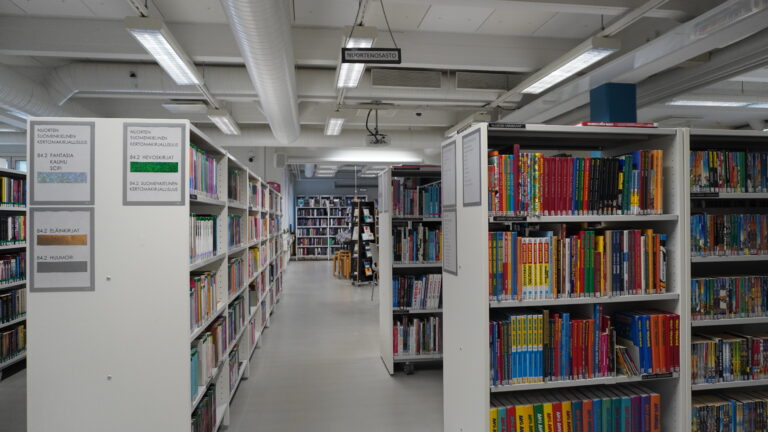 Kuvassa on kirjastosalin hyllyjä täynnä kirjoja